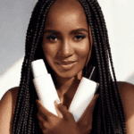 Person with long box braids holding two hair care product bottles against a softly lit background.